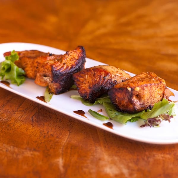 Four pieces of Indian-style grilled salmon with greens and sauce on a white rectangular plate.